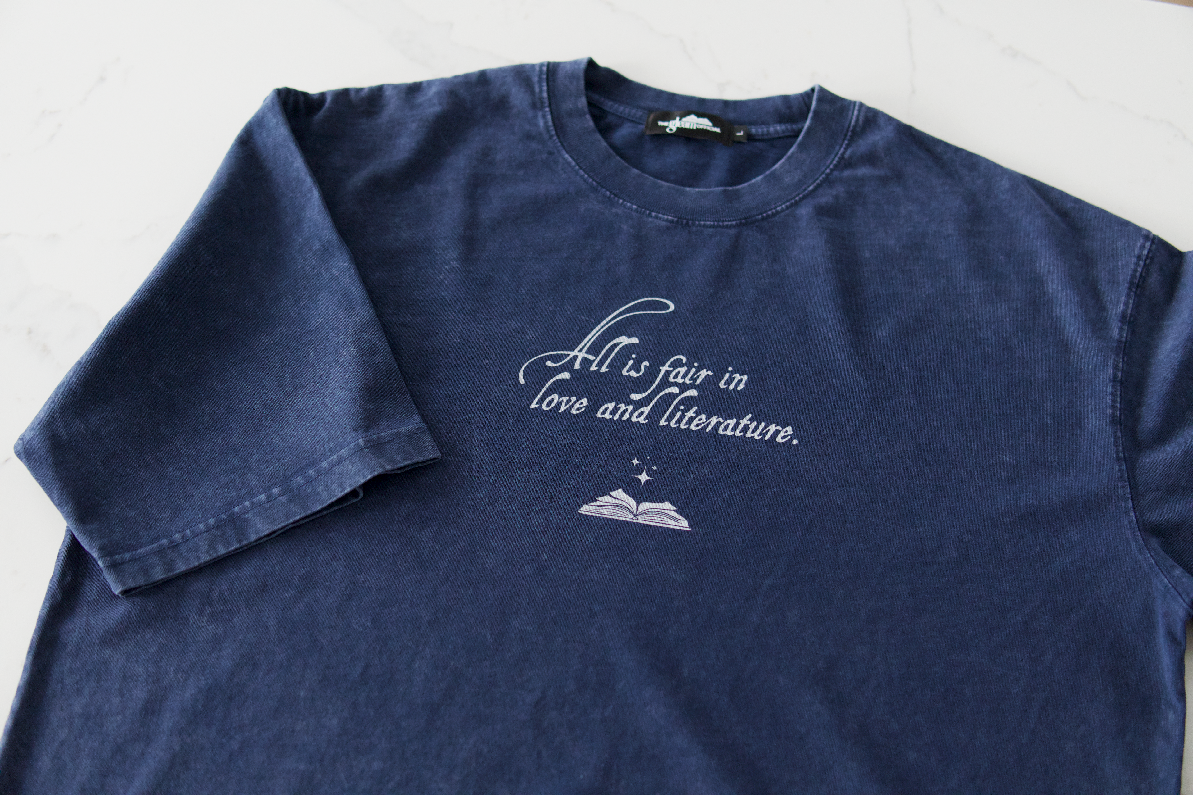 Blue mineral wash boxy cotton t-shirt with “All Is Fair in Love & Literature” text design laid flat