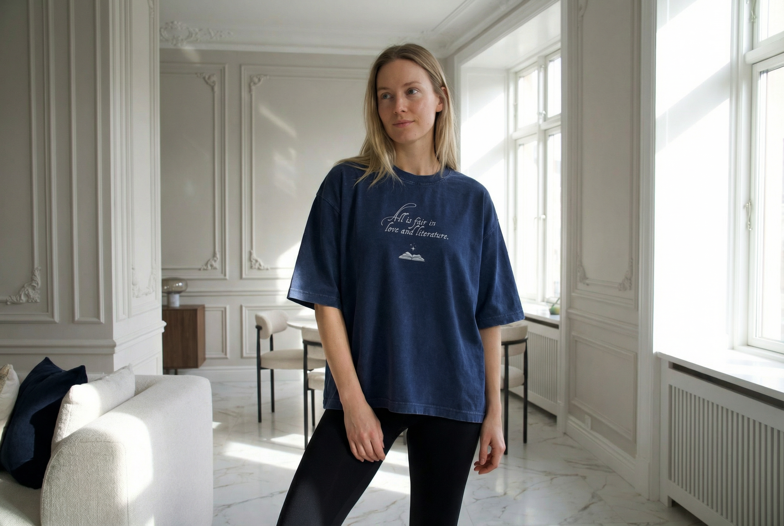 Model wearing a navy boxy t-shirt with “All Is Fair in Love & Literature” text, styled in a light, elegant neoclassical interior, designed for book lovers and readers