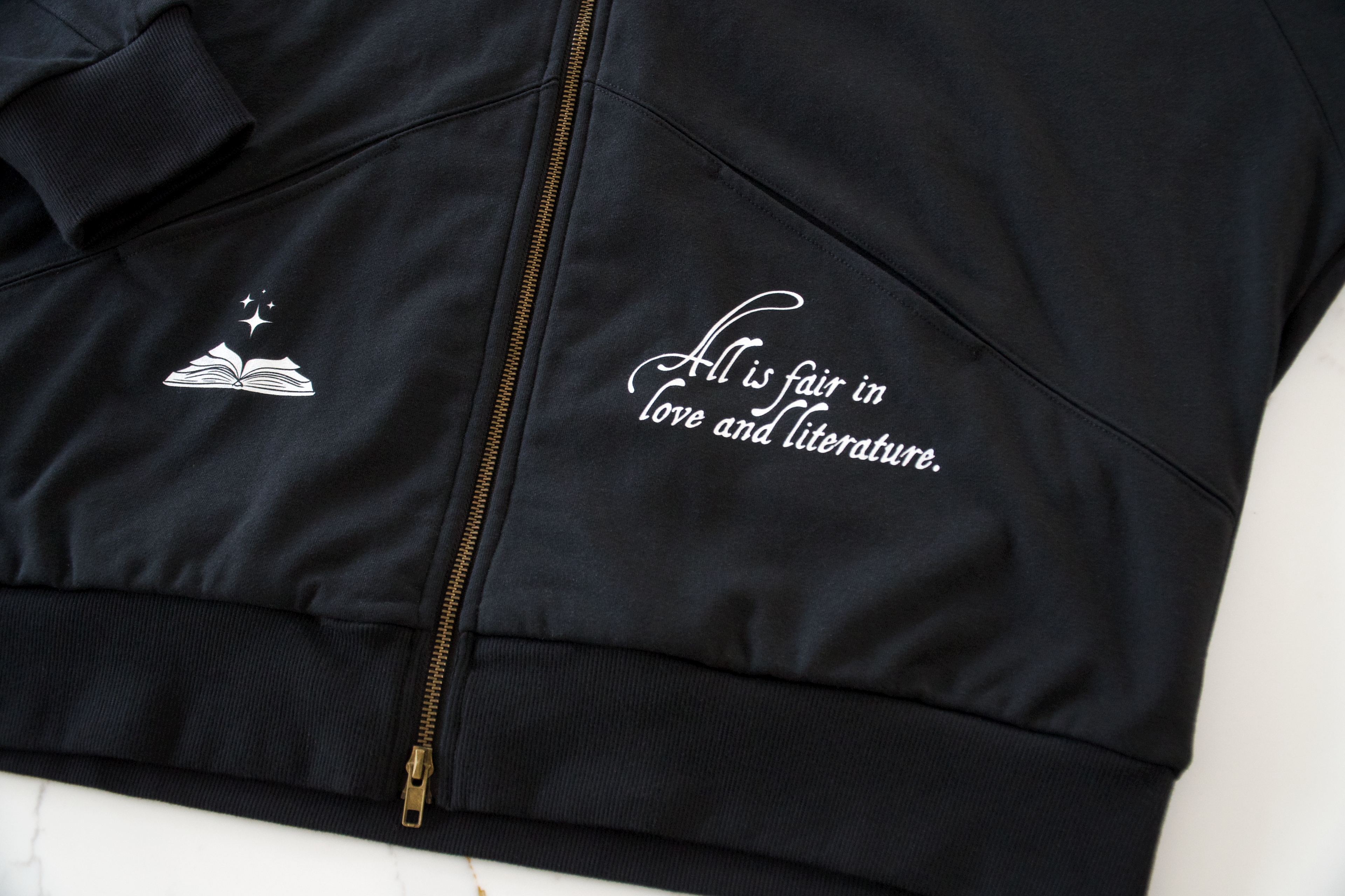 Flat lay close-up of black zip hoodie showing gold zipper and “All Is Fair in Love & Literature” chest design
