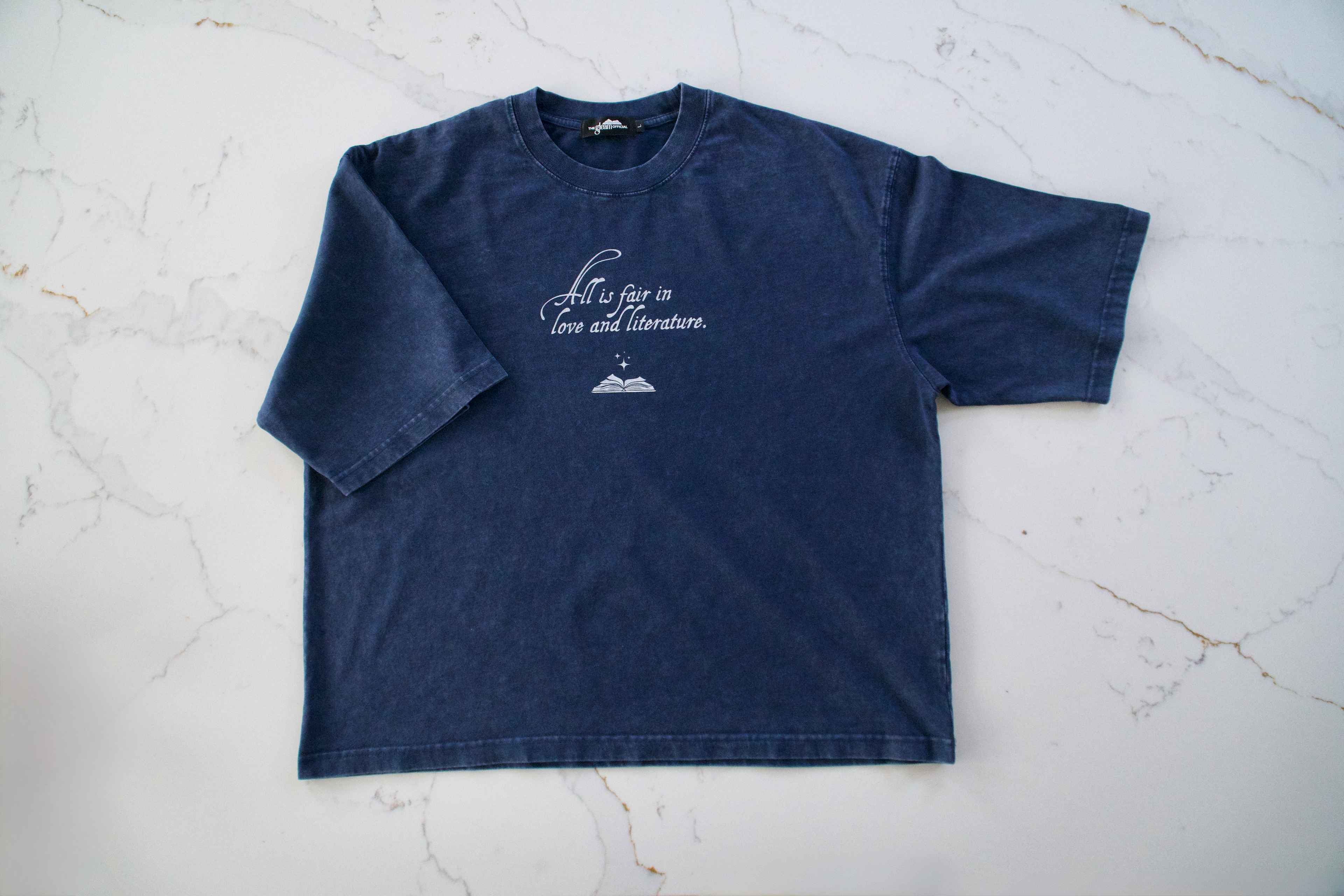 Blue mineral wash boxy cotton t-shirt with “All Is Fair in Love & Literature” text design laid flat