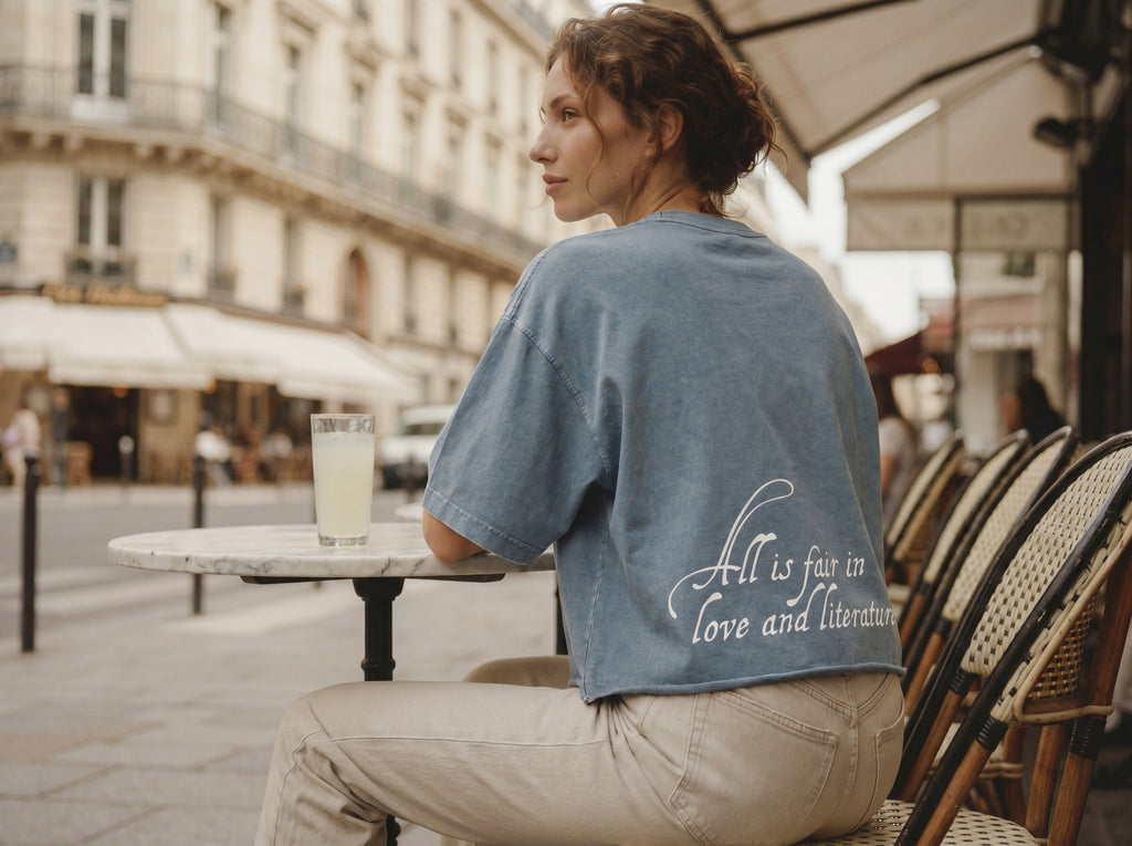 Woman sitting at an outdoor cafe table with a quote that reads "All is fair in love and literature" on her shirt.