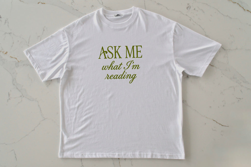 White t-shirt with 'Ask Me What I'm Reading' text on a light marble background