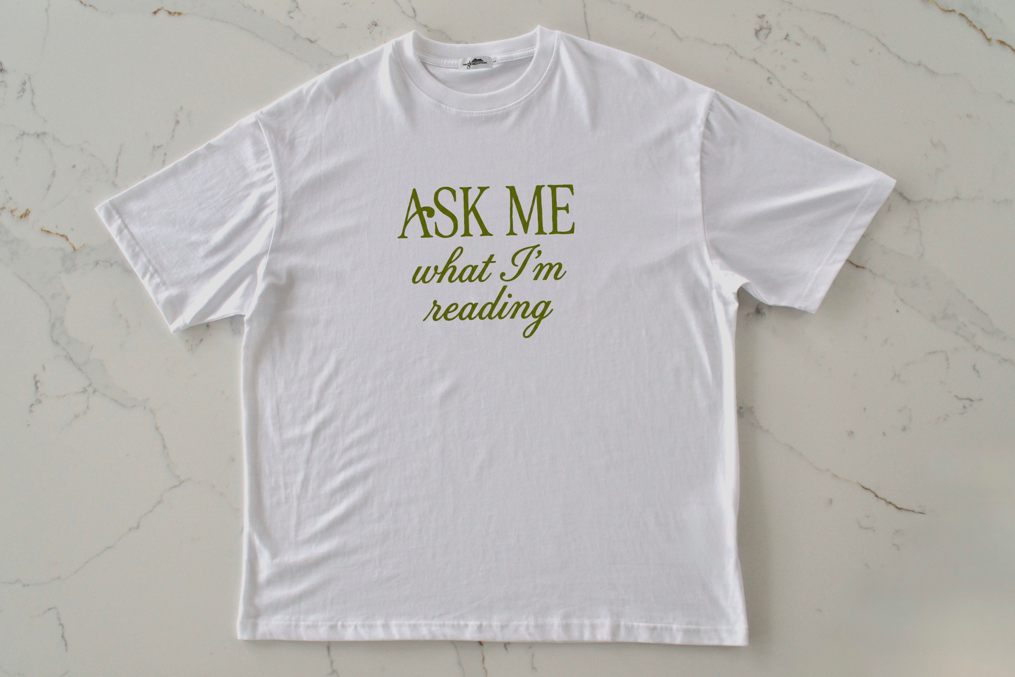 White t-shirt with 'Ask Me What I'm Reading' text on a light marble background