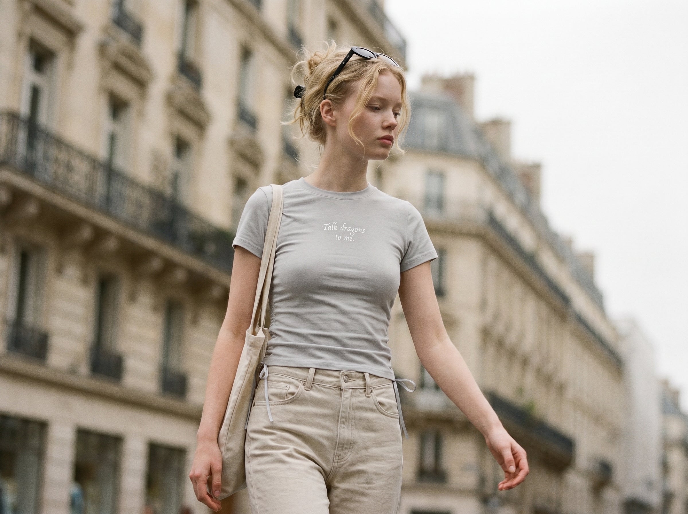 Woman walking on a city street wearing a gray t-shirt and beige pants. Shirt reads "talk dragons to me."