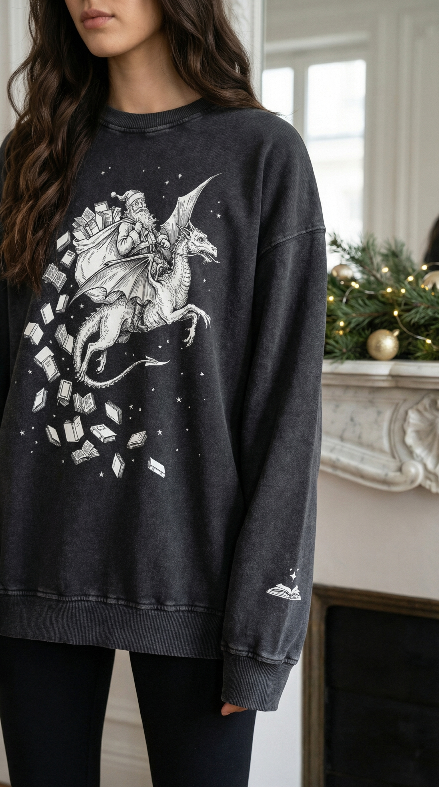 A woman wearing a charcoal-grey sweatshirt featuring a detailed white illustration of Santa riding a dragon, with books flying around them and small stars scattered across the design. A small white open-book graphic appears on the left sleeve. She is standing in a decorated room with greenery and gold ornaments on a mantel behind her.