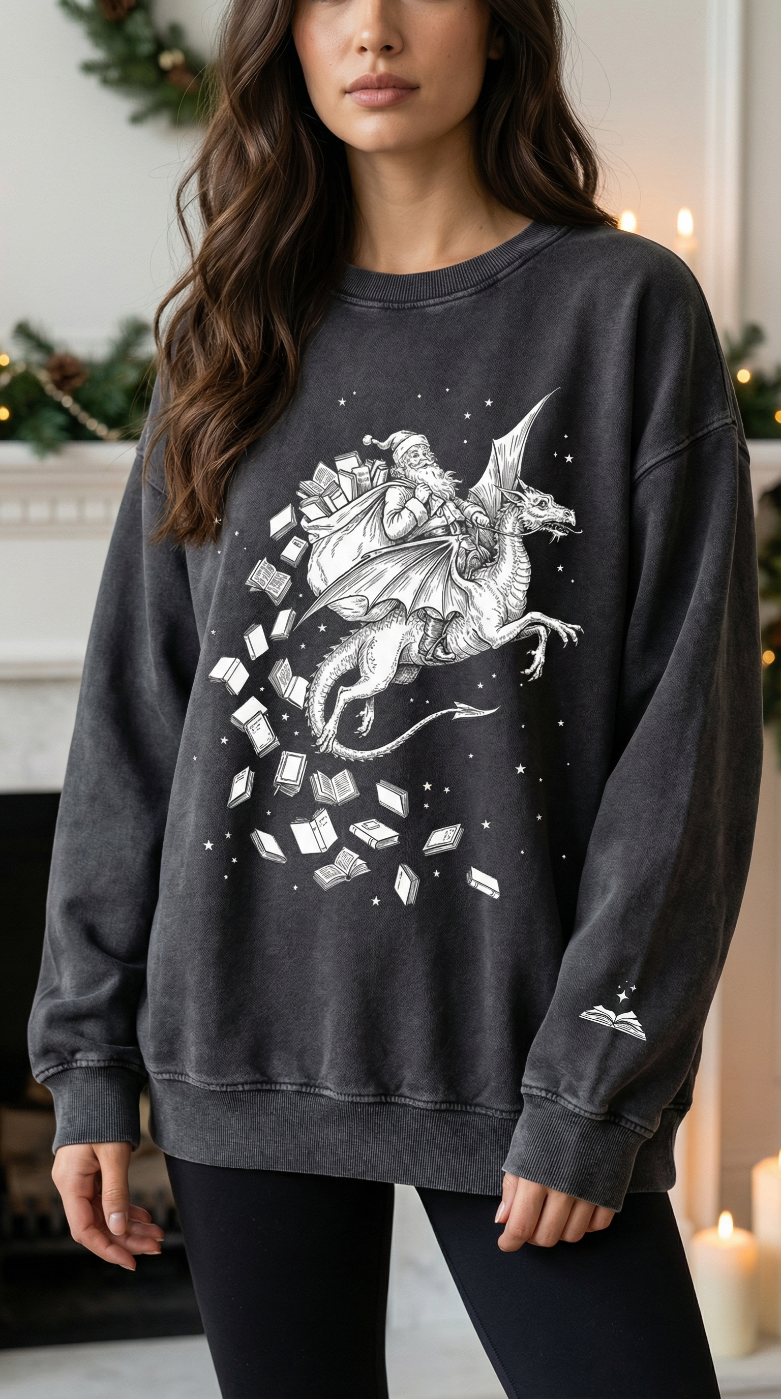 A woman wearing a charcoal-grey sweatshirt with a large white illustration on the front depicting Santa riding a dragon, surrounded by scattered flying books and small stars. A small white open-book graphic is printed on the left sleeve. She stands indoors in front of holiday decorations, including greenery and lit candles.