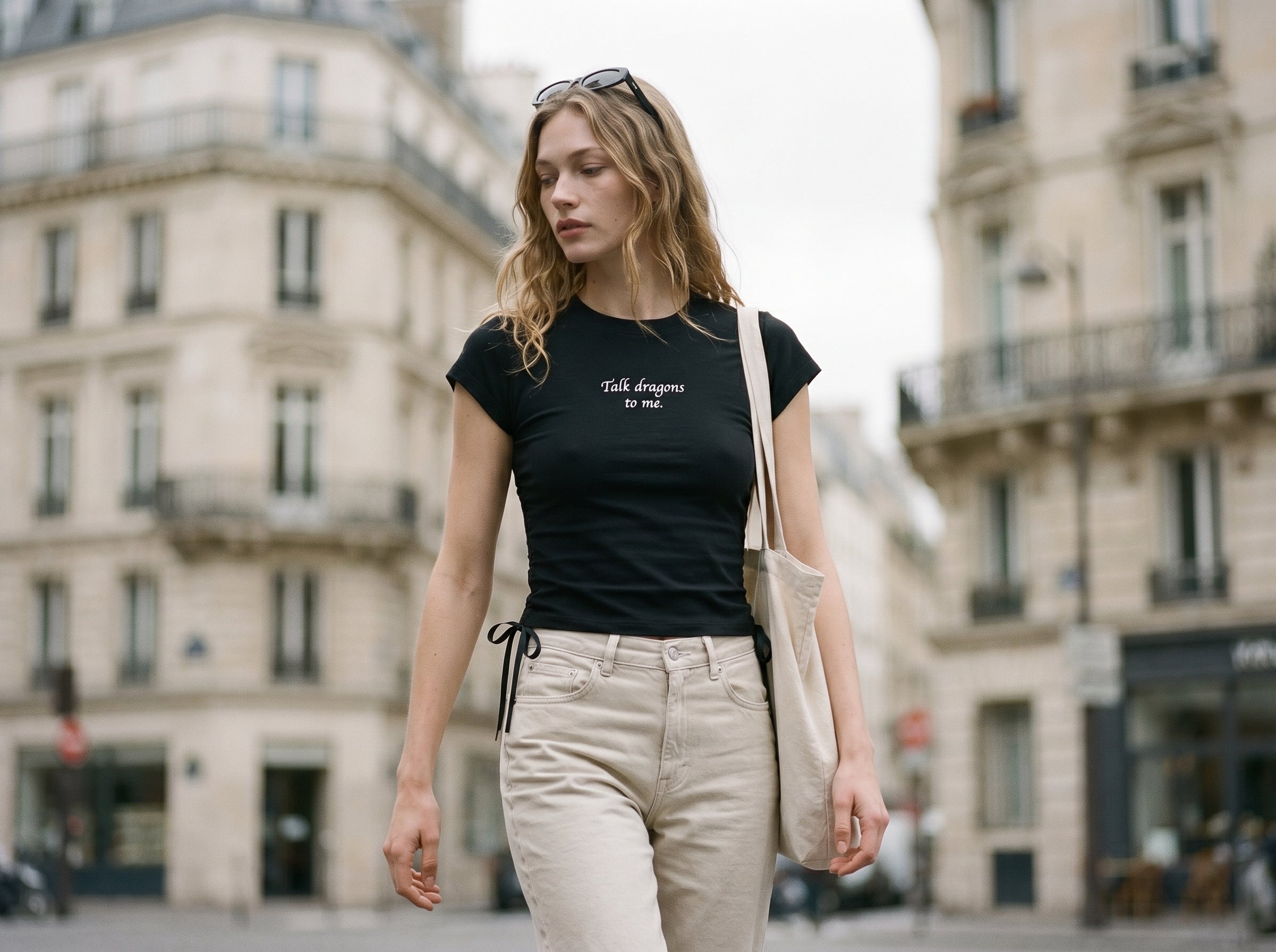 Woman walking on a city street wearing a black t-shirt with text and beige pants. Shirt reads "Talk dragons to me."