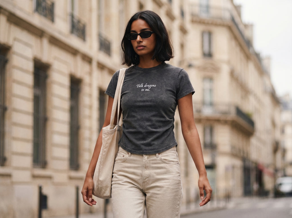 Woman walking on a city street wearing a charcoal gray t-shirt with text that reads "talk dragons to me" and light-colored pants.
