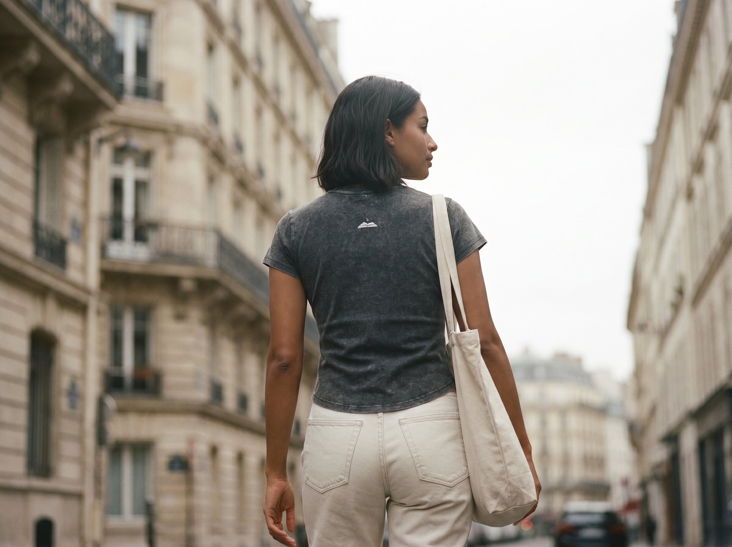 Woman walking down a city street holding a beige tote bag. Charcoal grey t shirt with a gleam logo on the back 