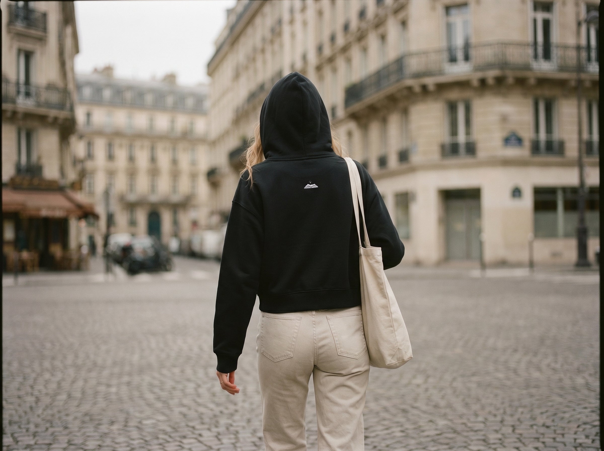 Person wearing a black hoodie with a gleam logo walking on a cobblestone street in an urban setting.