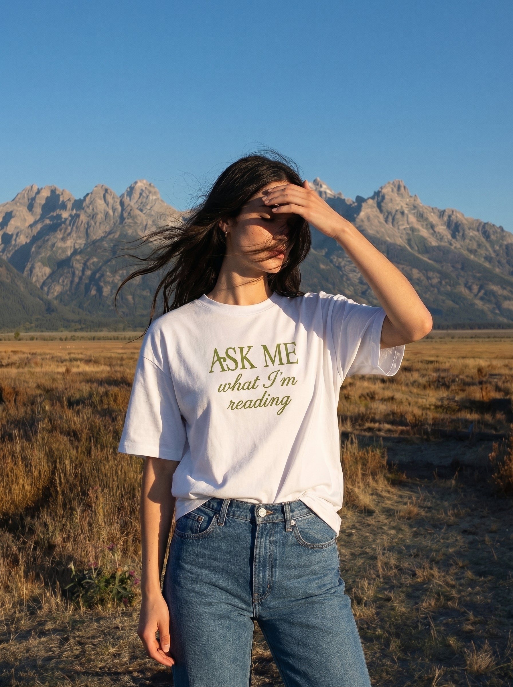 A young woman wearing a white t-shirt with 'Ask Me What I'm Reading' text in a desert landscape with mountains.