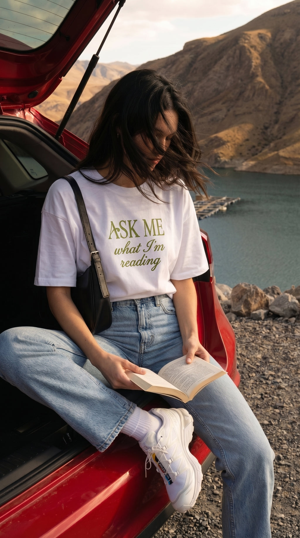 A young woman sitting on a car reading a book with mountains and lake in the background