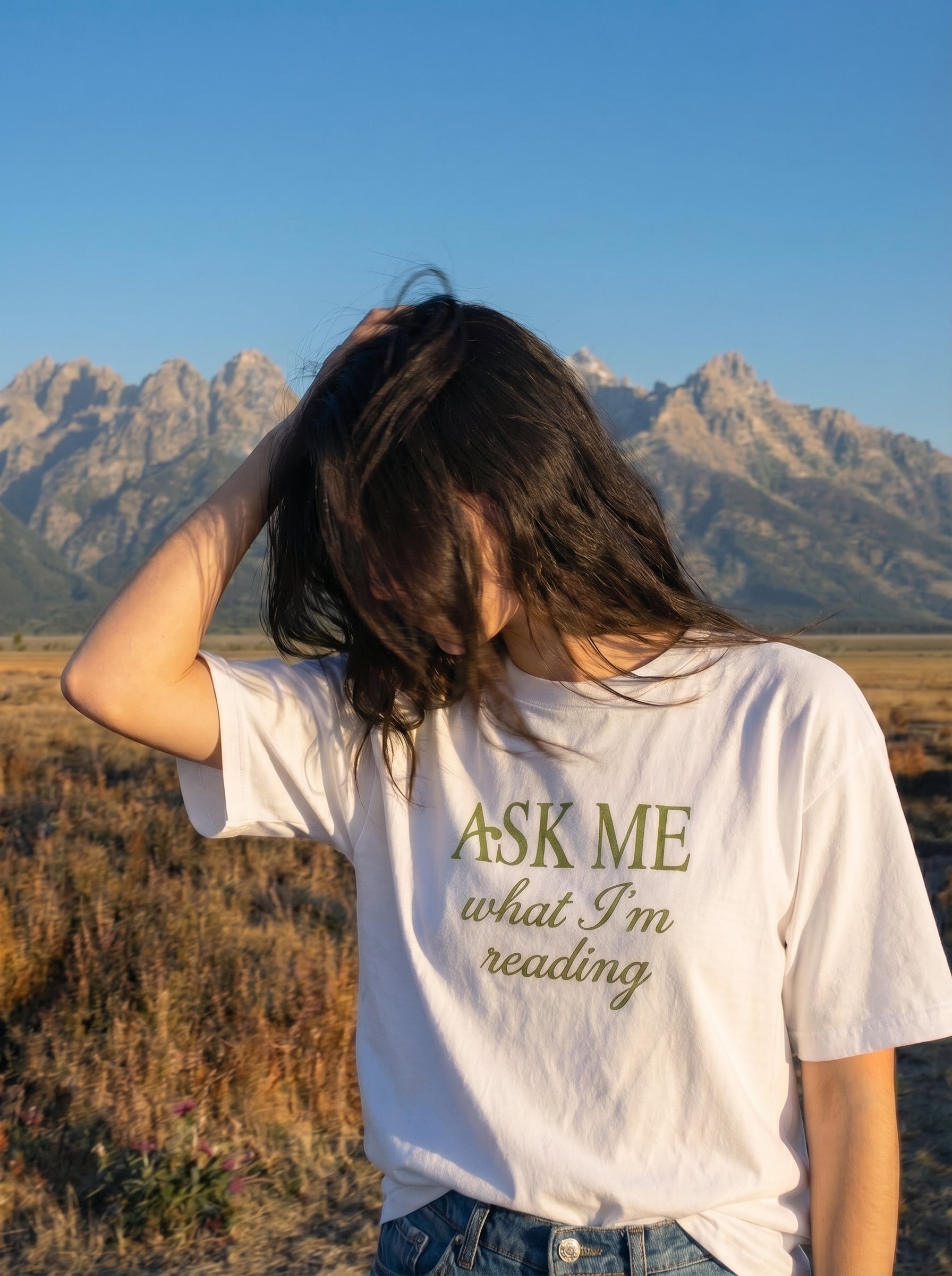 A young woman wearing a white t-shirt with 'Ask Me What I'm Reading' text in a desert landscape with mountains.